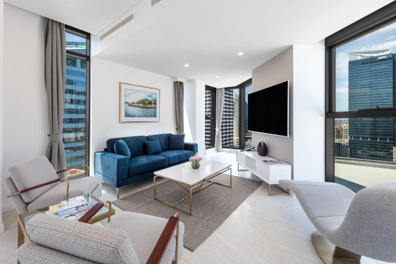 Meriton Suites Sussex Street, Sydney Buitenkant foto The photo shows a modern living room featuring large windows that allow plenty of natural light. The room is elegantly designed with a blue sofa, a light-colored coffee table, and two armchairs. There’s a flat-screen TV mounted on the wall, and a pie